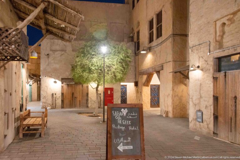 Nighttime view of a narrow stone-paved alley in Al Seef’s heritage district, lined with plaster buildings, benches, and shop entrances under wall lamps, with a freestanding sign in the foreground.