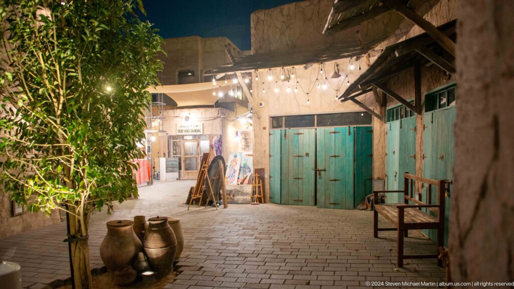 Traditional courtyard at Al Seef with bright turquoise double doors, rough plaster walls, rustic timber shade structures, and large clay jars beside a simple bench.