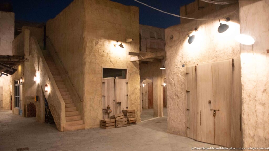 Traditional Al Seef courtyard with rough plaster walls, an external stair along the left wall, timber doors, hanging lamps, and simple seating.