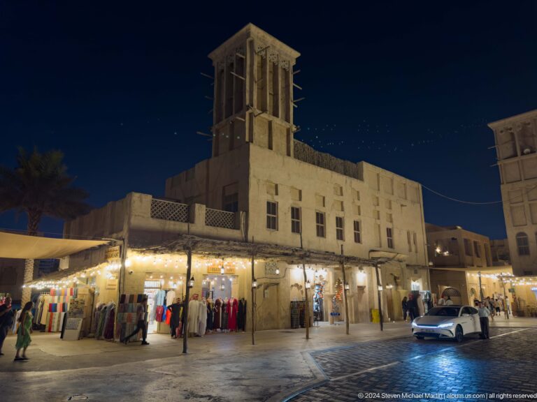 Nighttime view of a public square in Al Seef with pedestrians walking past illuminated shopfronts beneath a prominent square wind tower rising above low façades.