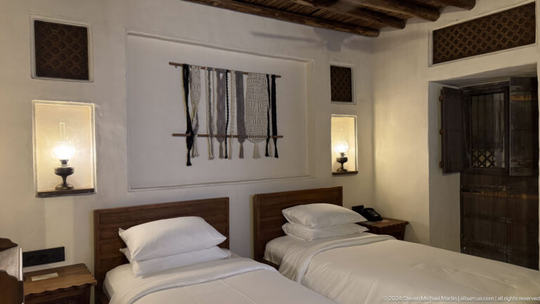 Twin hotel room at Al Seef in Dubai with two single beds beneath a dark vertical textile artwork, featuring neutral fabrics and dark wood details in a symmetrical heritage-inspired design.