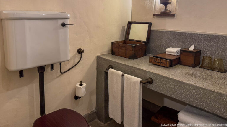 Heritage-style hotel bathroom vanity with a thick gray stone countertop, folded white towels, tissue box, toiletries, and a framed mirror above wall-mounted fixtures.
