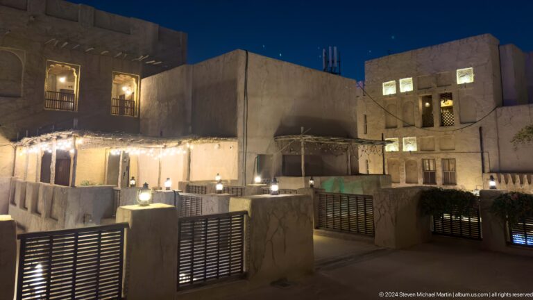 Nighttime elevated view across Al Seef’s flat, parapeted rooftops and cream plaster low-rise buildings lit by warm exterior lamps against a dark sky.