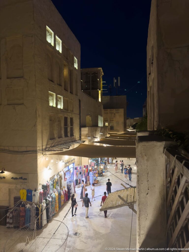 Nighttime elevated view of a narrow market street at Al Seef in Dubai, with pedestrians moving along a brightly lit lane between darker upper terraces.