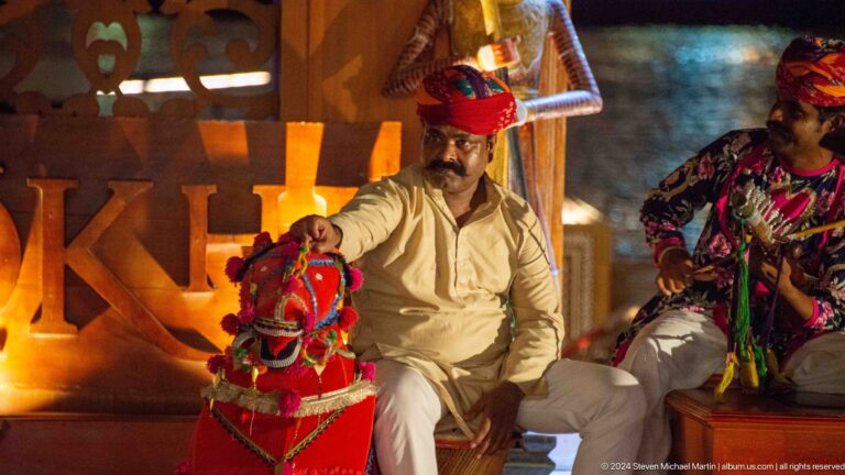 Man in white robe and red head covering seated beside large colorful puppet figures under warm stage lighting at Al Seef in Dubai.