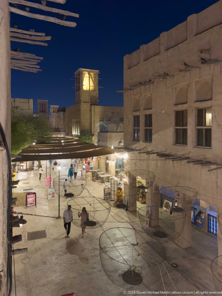 Nighttime view from above of a crowded pedestrian shopping street in Al Seef, lined with brightly lit shops and traditional-style plaster buildings.