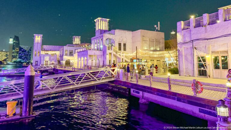 Night view of the Al Seef waterfront in Dubai with a low metal pedestrian bridge crossing reflective water in front of illuminated wind-tower buildings.