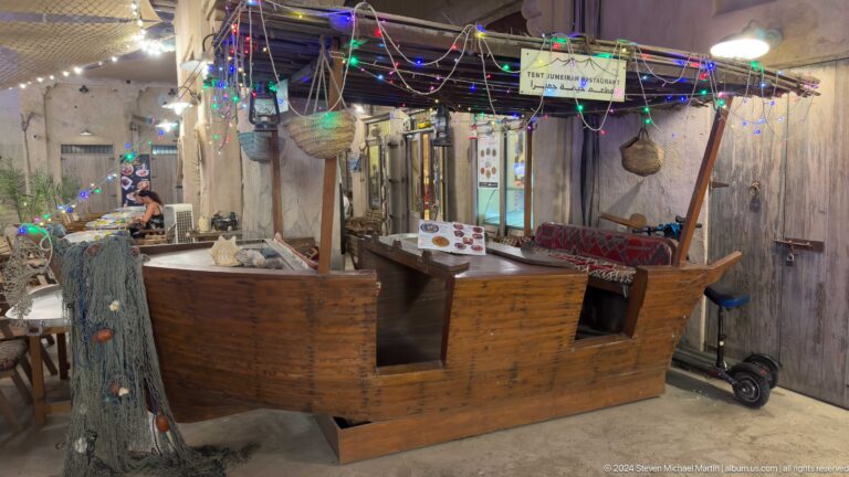 Old wooden cart-like stall positioned against a rough plaster wall under hanging colored lights in an indoor market-style space.