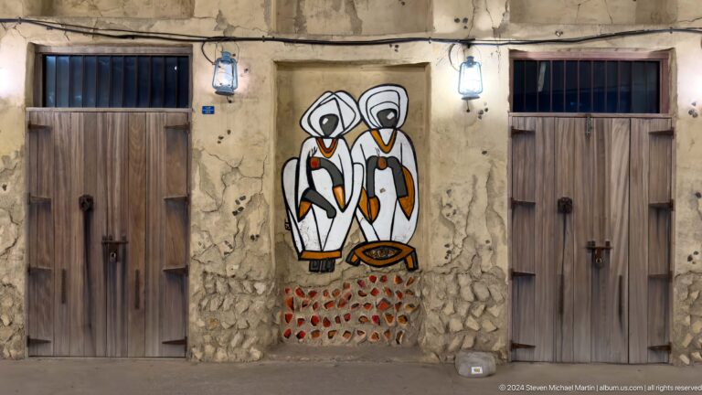 Stylized mural of three Gulf figures in white head coverings mounted between wooden doors on a rough plaster wall in Al Seef, Dubai.