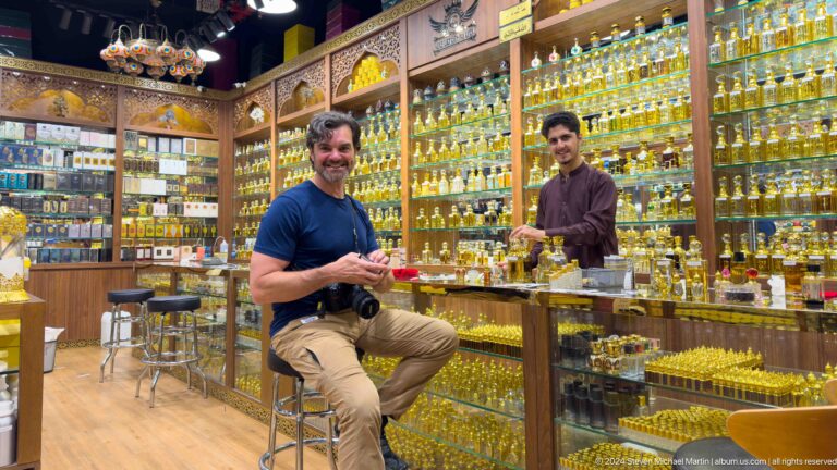 Customer and shopkeeper seated facing the camera in a perfume shop at Al Seef, with rows of glass fragrance bottles filling the background.
