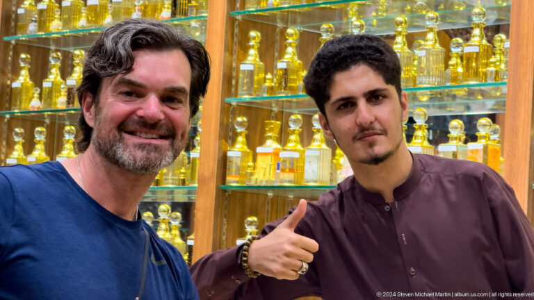 Close portrait of a visitor and perfume seller giving a thumbs-up in front of brightly lit shelves filled with glass perfume bottles at Al Seef in Dubai.