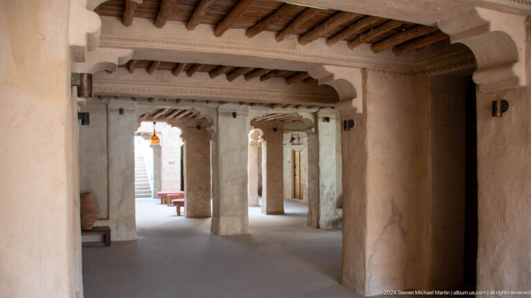 Daytime view along a heritage-style covered corridor in Al Seef, Dubai, with thick plaster columns, exposed timber ceiling beams, benches, hanging lamps, and bright light at the far end.