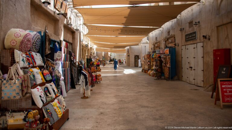 Daytime view of a broad shaded market corridor near Al Seef and Al Fahidi in Dubai, with textiles and souvenirs displayed along storefronts on both sides under a continuous canopy.