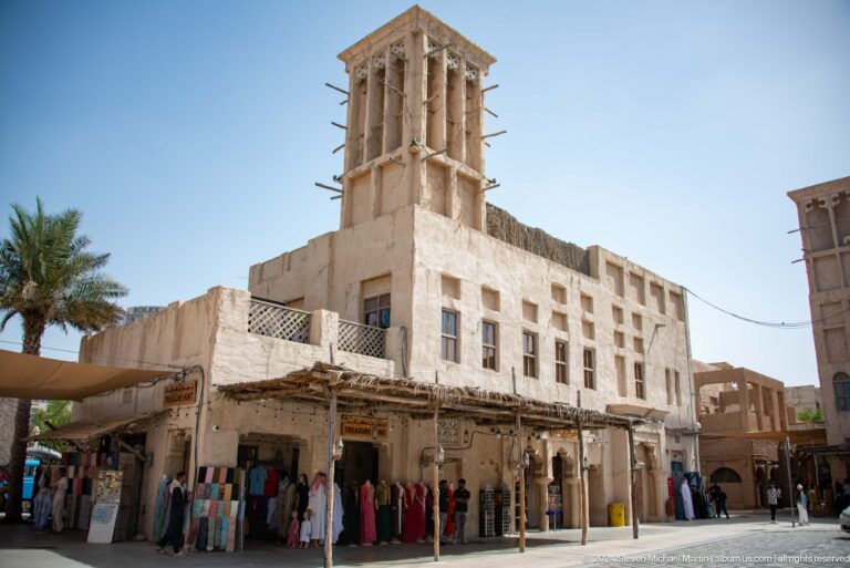 Square traditional wind tower atop a cream plaster heritage building with shaded ground-floor frontage in the Al Fahidi Historical Neighbourhood, Dubai.