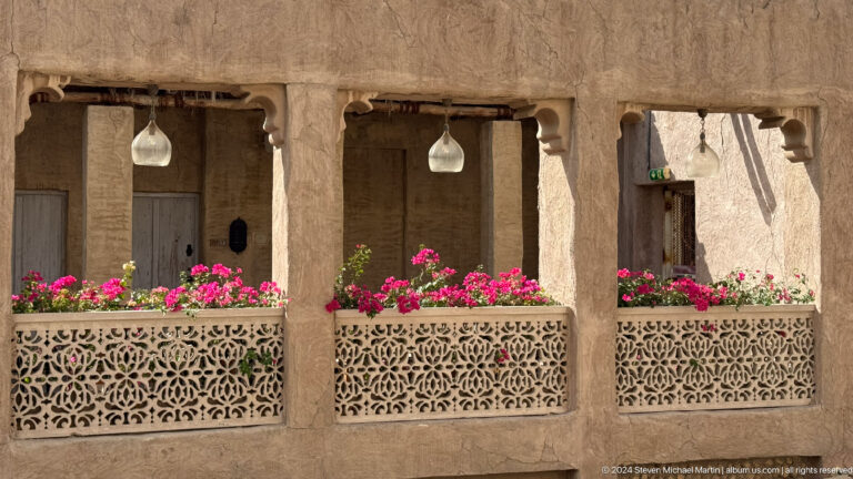 Earth-toned plaster façade in Al Fahidi, Dubai, with three dark timber windows and an ornate projecting wooden balcony decorated with pink flowers.