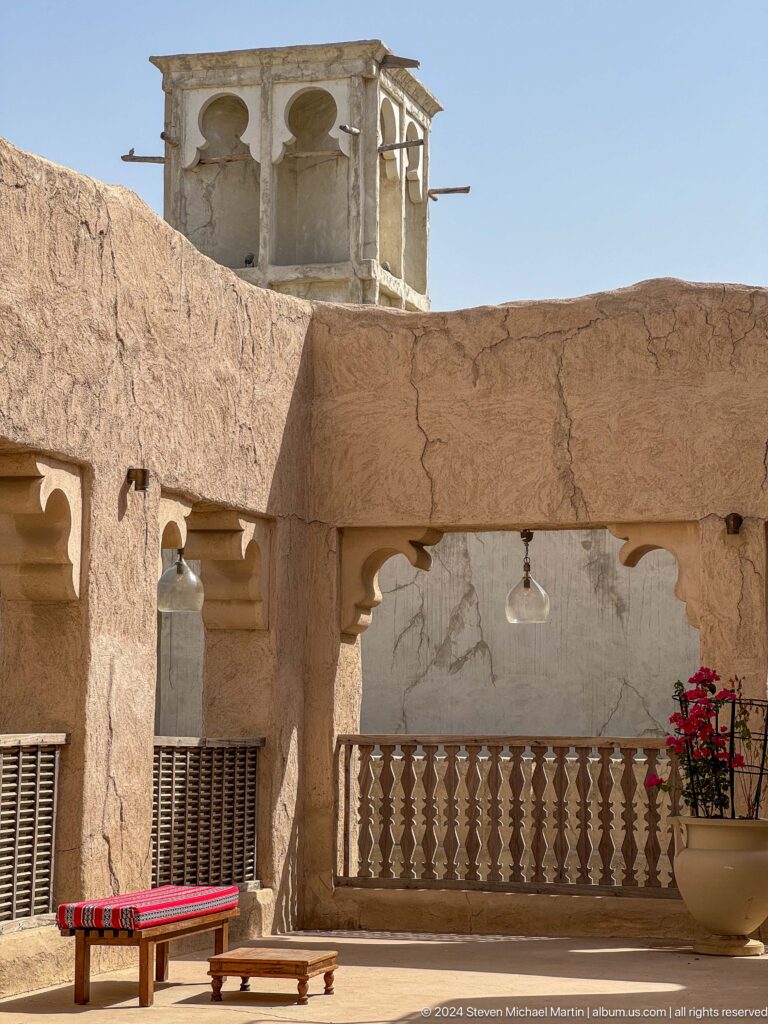 Shaded arcade with thick plaster arches and columns surrounding a small courtyard, with hanging lamps and a flower arrangement in Al Fahidi.