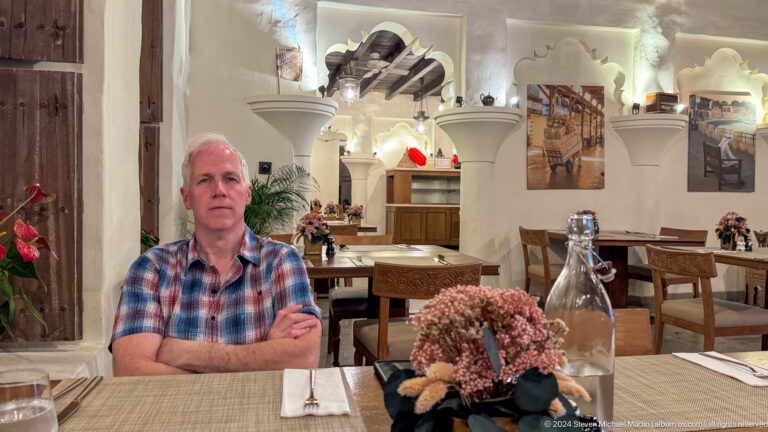 Gray-haired man in a plaid shirt sits at a dining table with flowers and a glass bottle in a heritage-style Dubai restaurant with white arches and rustic wall decor in the background.