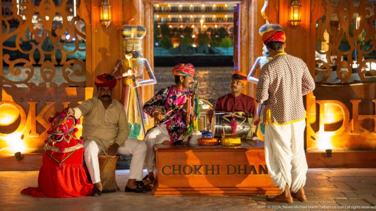 Two men in white traditional clothing stand near a colorful heritage stage with a large red seated figure and multicolored theatrical props in Al Seef, Dubai.