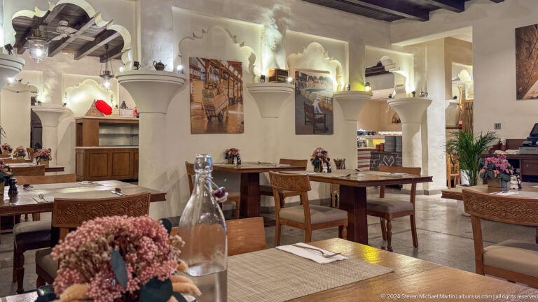 Empty heritage-style restaurant dining room in Al Fahidi, Dubai, with wooden tables and chairs arranged between white arches and columns.
