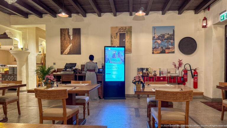 Heritage-themed restaurant dining room in Dubai’s Al Fahidi area, with wooden tables and chairs facing a tall vertical digital display under exposed beams.