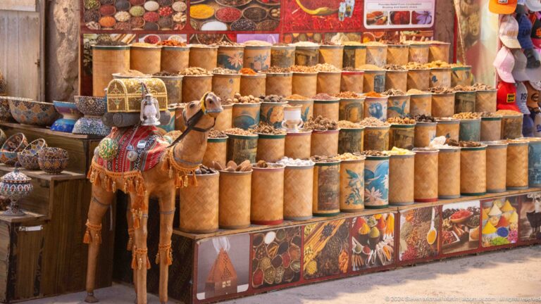 Open-air market stall with tall cylindrical containers filled with assorted spices and dried goods in shades of brown, red, orange, and beige, arranged in rows in Dubai.