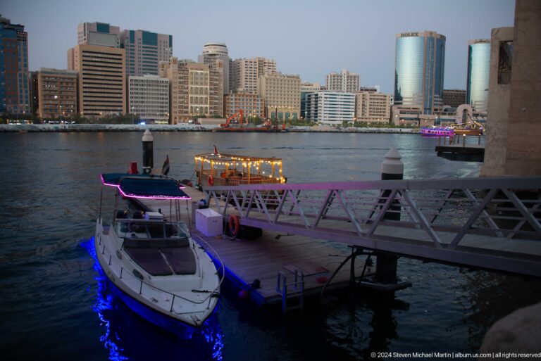 Illuminated multi-level cruise boat moored at an Al Seef dock on Dubai Creek at night, with city buildings visible across the water.