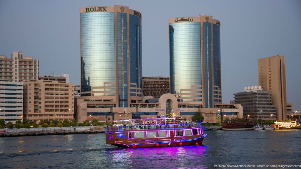 Purple-lit sightseeing boat crossing Dubai Creek at night with twin blue-glass towers in the background.