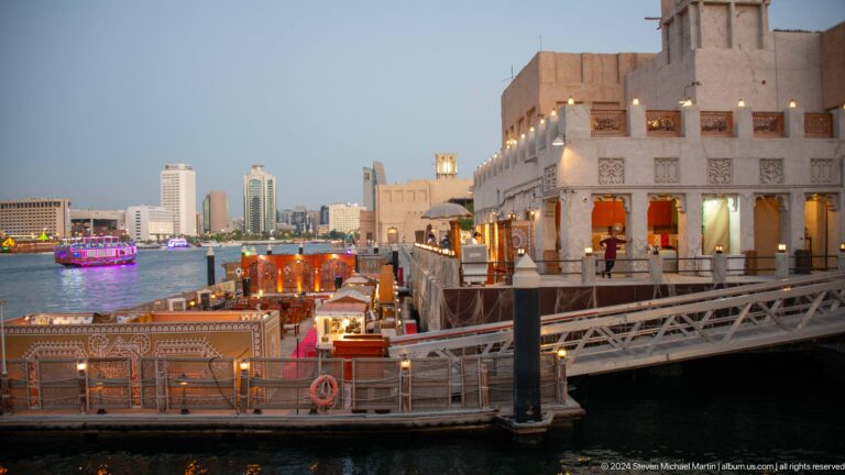 Wooden passenger boat passing cream-colored heritage-style buildings along the Al Seef waterfront promenade on Dubai Creek.