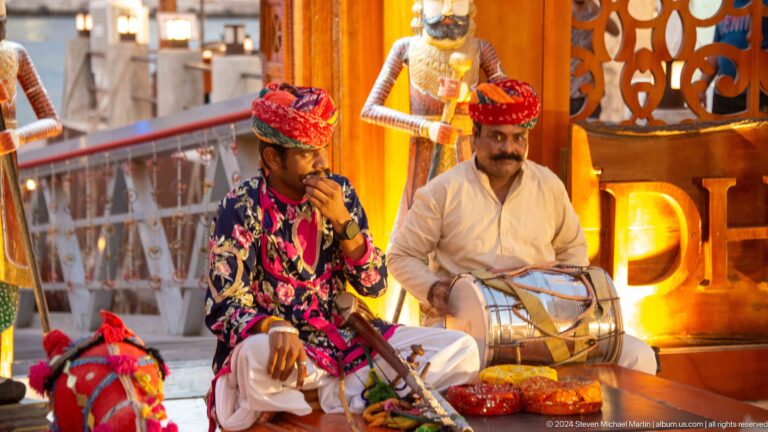 Two men in white garments and red head coverings sit beside bright red and multicolored decorative figures during a cultural performance at Al Seef.