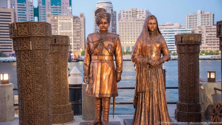 Life-size bronze sculptures of a man and woman in traditional Gulf dress installed beside a carved architectural screen in an outdoor setting.
