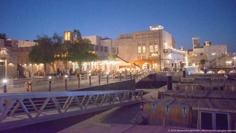 Evening view of Al Seef’s low-rise, illuminated waterfront buildings reflected in the calm creek beside a bridge and railing under a deep blue sky.