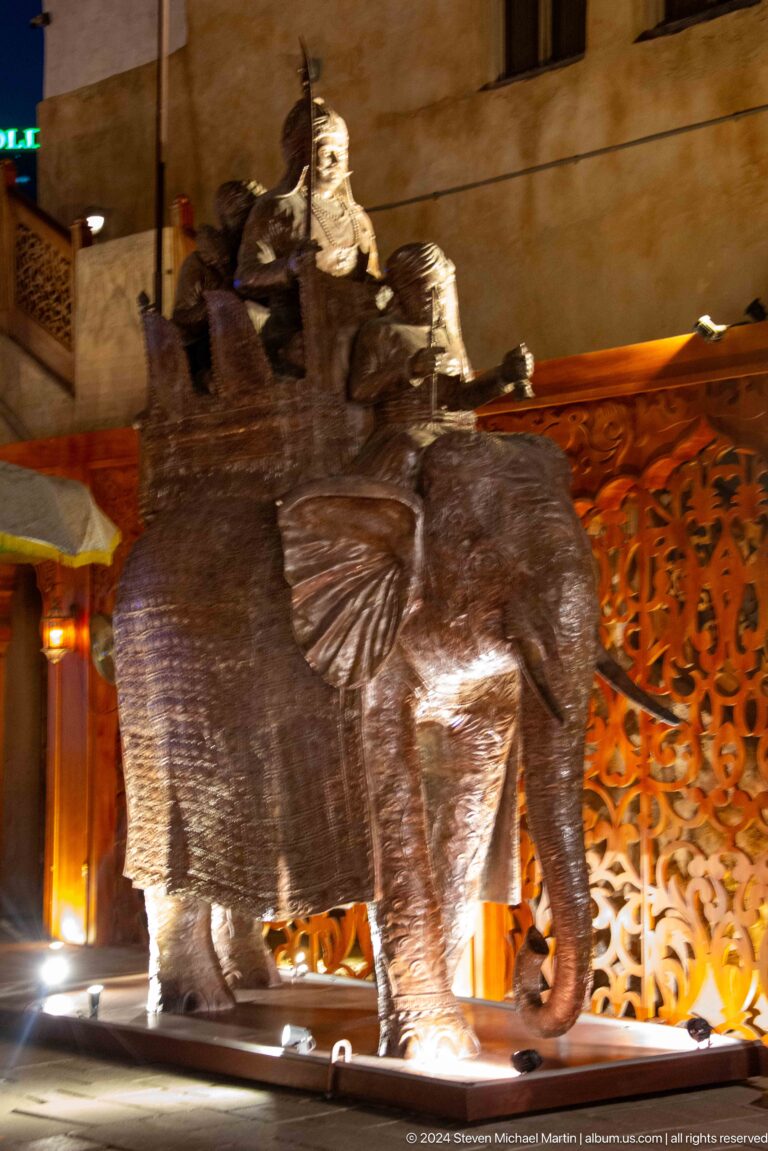 Close-up of a dark decorative elephant sculpture with raised trunk set before an intricately carved wooden panel.
