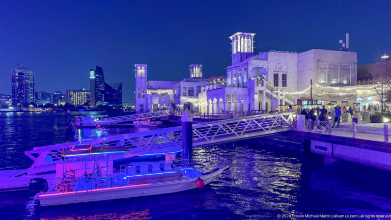 Night view of a pedestrian bridge at Al Seef in Dubai, with purple and blue lighting reflected in the water and illuminated heritage-style buildings with wind towers in the background.