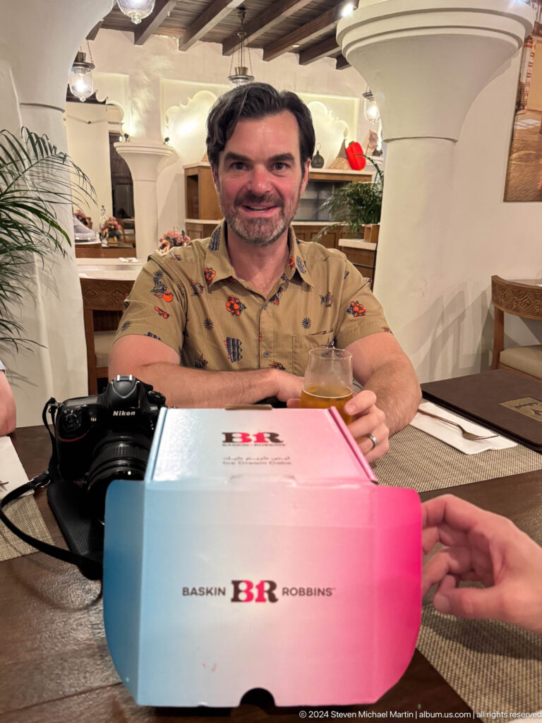Smiling man in a patterned shirt seated at a restaurant table in Al Seef, Dubai, with a camera on the table as he holds a pink card before a birthday celebration.
