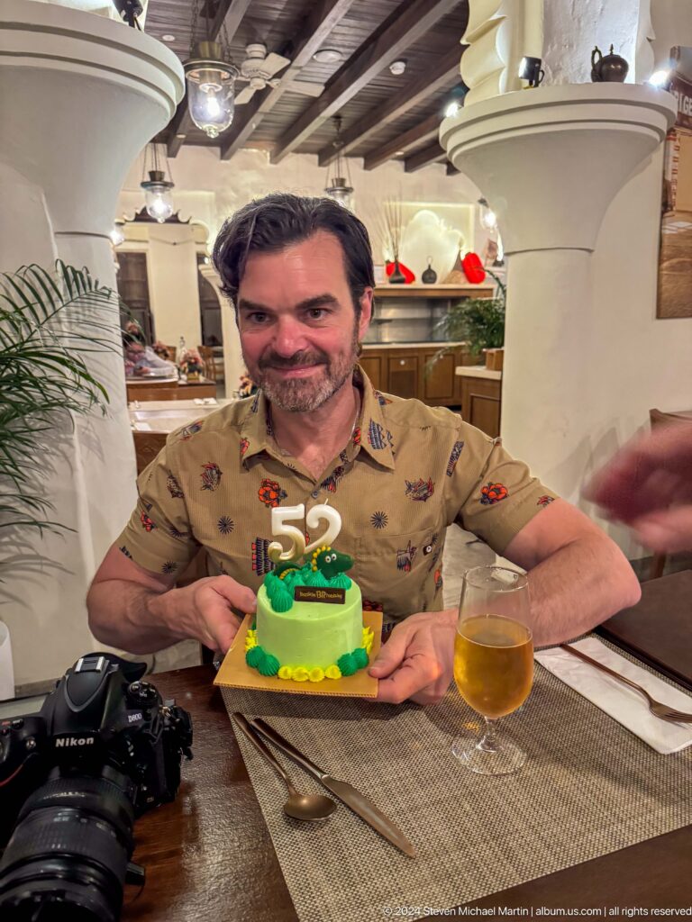 Man seated at a restaurant table smiling behind a small green cake with a large 52 topper and a drink beside him.