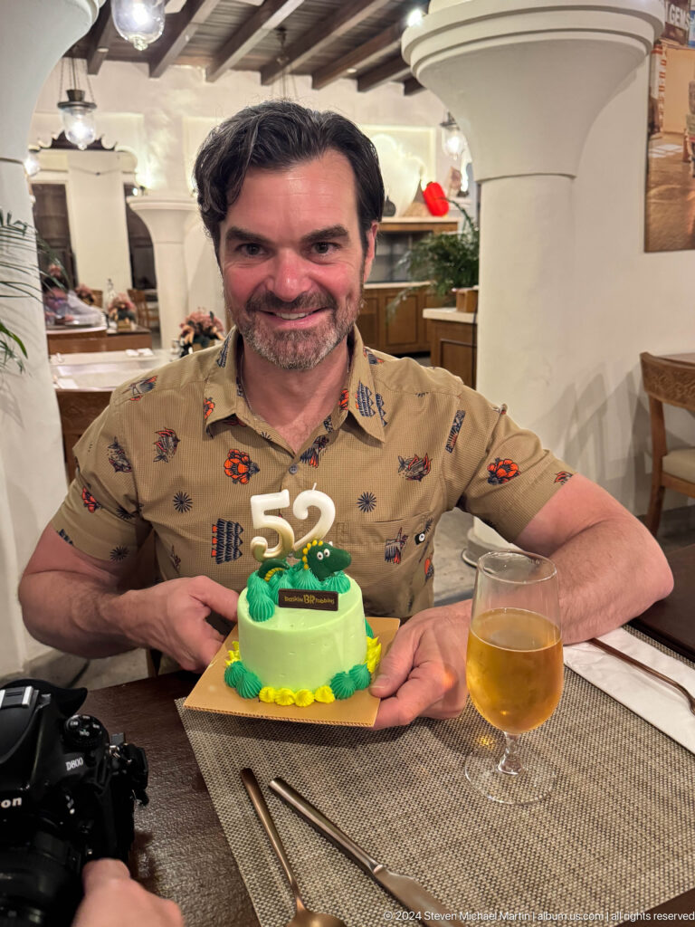 Smiling man in a patterned shirt leaning behind a green birthday cake with a large 52 topper and a glass of beer on a restaurant table.