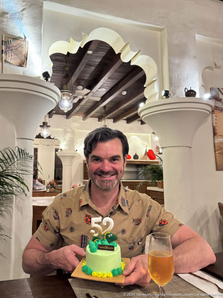 Smiling man in a patterned shirt seated in a heritage-style Al Seef restaurant in Dubai, with white columns and dark ceiling beams behind him.