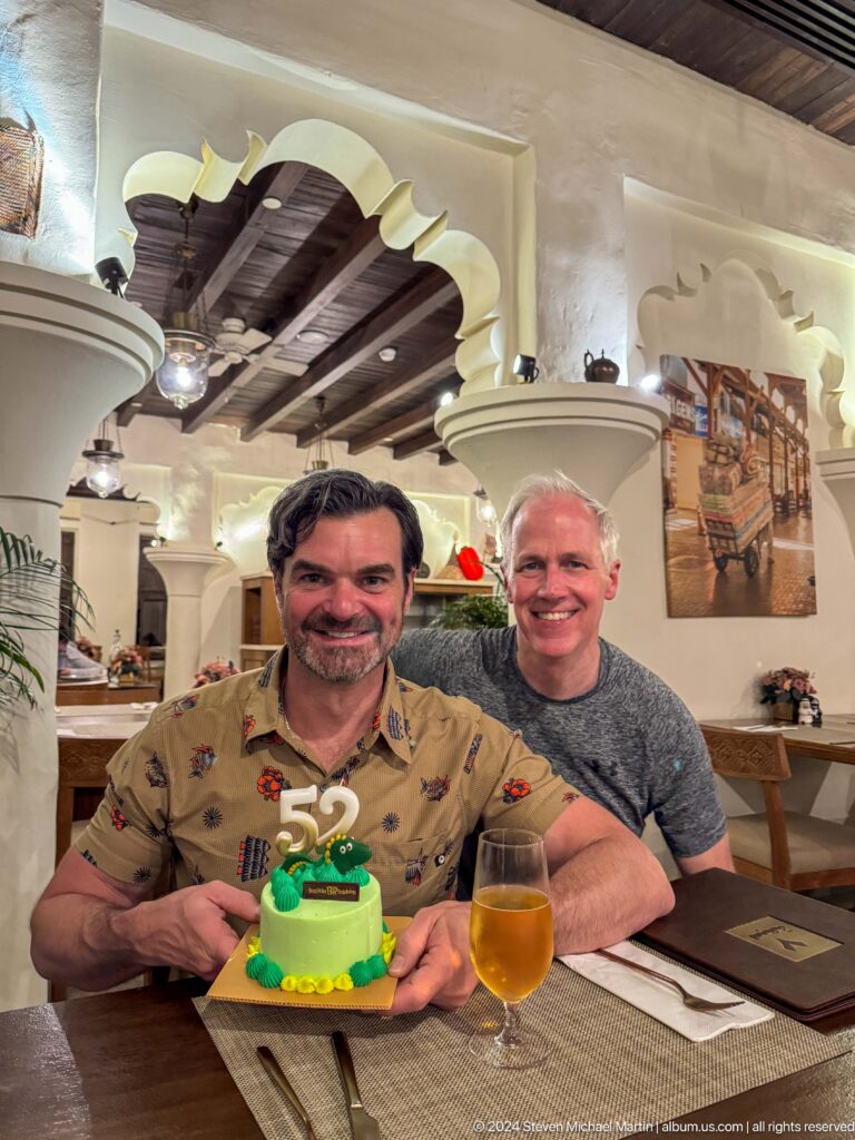 Two men lean together and smile for the camera in a restaurant at Al Seef, Dubai, with white arches and wall art behind them.