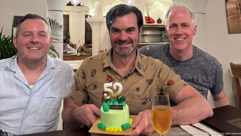 Three men smiling at a restaurant table with a small green birthday cake topped with the number 52 in front of them.