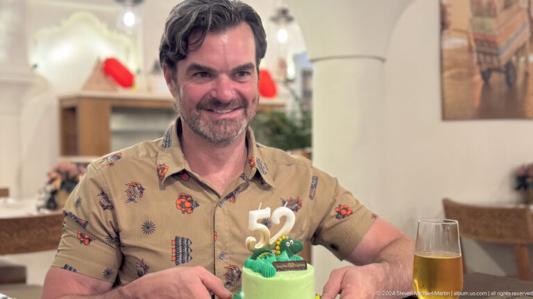 Man seated at a restaurant table during a birthday dinner, with a green cake and drink in front of him, looking toward the camera in a candid moment.