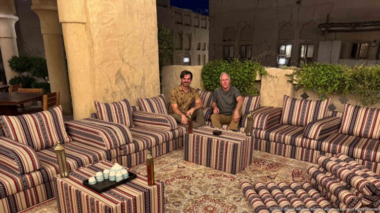 Two men sit on striped low majlis-style sofas with cushions and small tables in an outdoor lounge at Al Seef in Dubai.