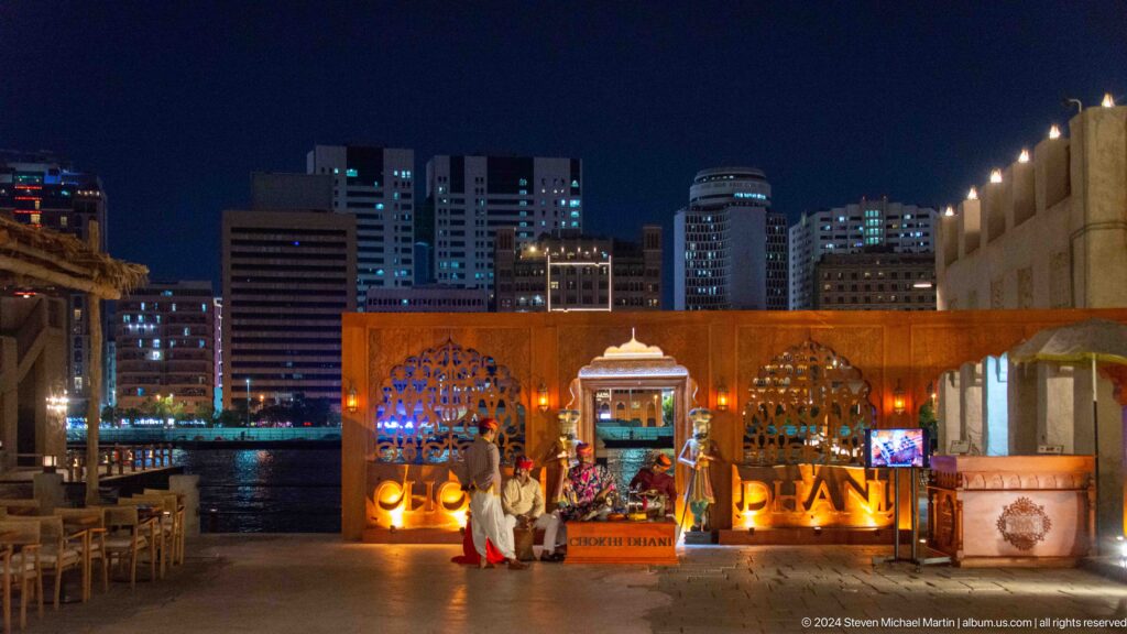 Night view of a lit waterfront stage at Al Seef with Dubai Creek in the foreground and modern high-rise buildings illuminated across the water.