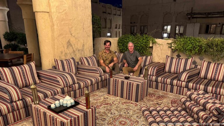 Wide view of two men seated on low striped, U-shaped sofas in a majlis-style lounge at Al Seef Dubai, framed by patterned cushions, small decorative tables, and foliage.