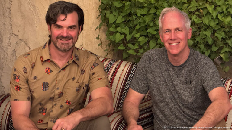 Two smiling men sit close together on striped cushions with green foliage behind them in a majlis-style seating area.