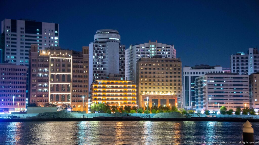 Night view of illuminated mid-rise and high-rise buildings across Dubai Creek with long reflections on the dark water.