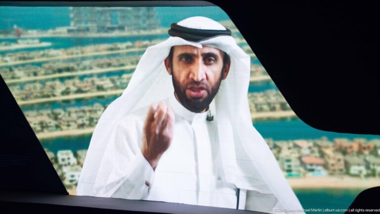 Emirati man in white traditional dress speaking on a large exhibition screen.