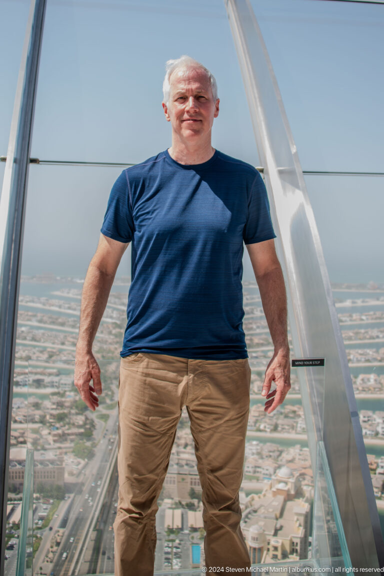 Close crop of a man in a navy shirt standing on a glass observation deck above Palm Jumeirah.