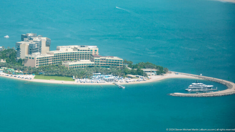 Low-rise Palm Jumeirah beach resort with loungers, turquoise water, and a yacht.