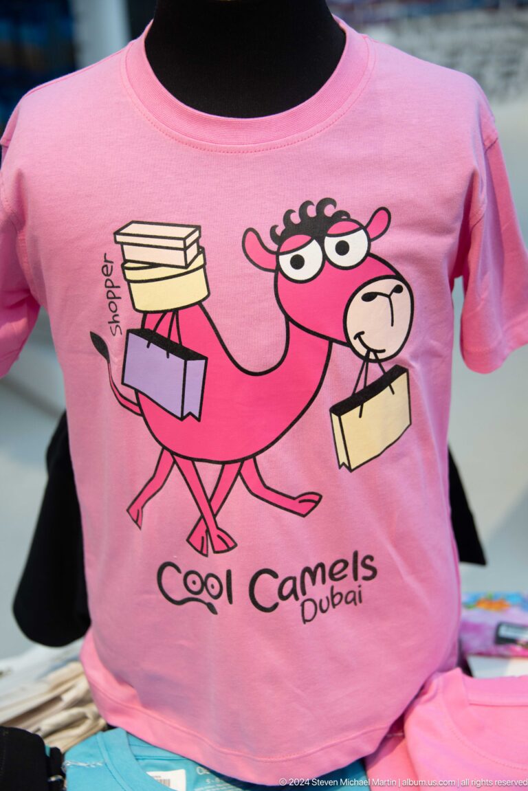 Pink souvenir T-shirt printed with a cartoon camel carrying shopping bags.
