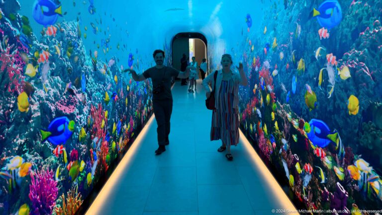 Two visitors walking through a bright blue corridor covered with tropical fish and coral.